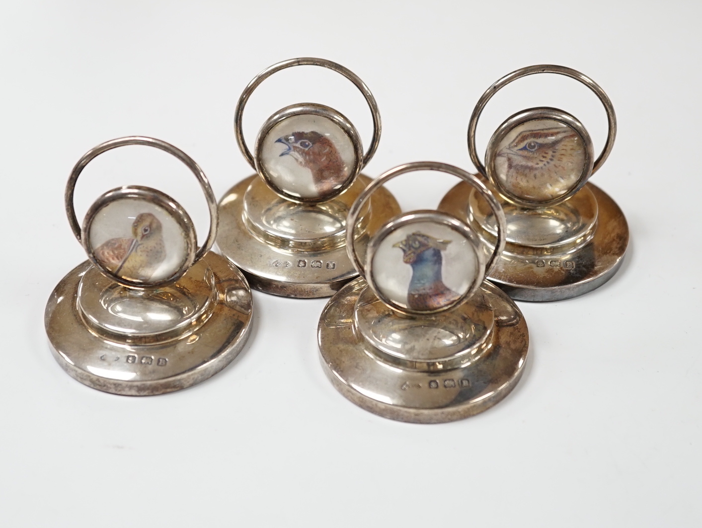 A set of four George V silver menu holders, each with inset painted panel of a wild bird, Adie Bros. Ltd, Birmingham, 1926, base diameter 5cm.